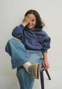 Sudadera con capucha azul marino y logotipo rosa, de corte relajado, combinada con jeans de pierna ancha en azul claro y calzado beige, sentada en una silla de madera.