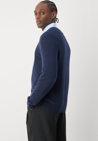 Un pull bleu marine en tissu tricoté doux avec un col rond, des poignets côtelés et un ourlet côtelé. Associé à un pantalon noir et une chemise bleu clair en dessous.