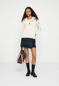 Casaco creme com fecho éclair e frente acolchoada, combinado com uma saia aos quadrados azul e verde, mocassins pretos e uma bolsa castanha. Tecido suave e design casual.