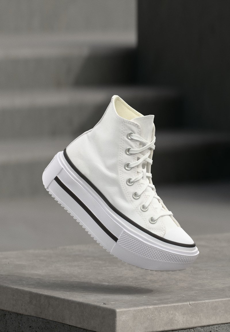 Sneaker alta bianca con suola spessa platform e striscia nera, sospesa sopra uno scalino di cemento all'aperto.