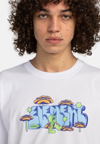 Witte katoenen t-shirt met kleurrijke graphics van bloemen en een cartoonfiguur. Het ontwerp bevat felle blauwe en oranje accenten.