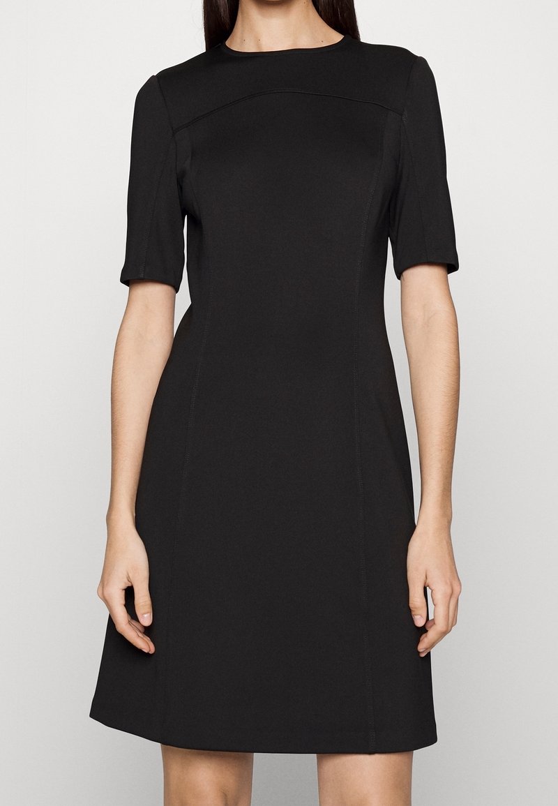 Femme portant une robe noire à manches courtes avec une taille cintrée et une jupe légèrement évasée, debout devant un fond uni.