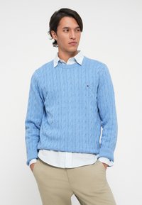 Young man wearing a light blue cable-knit sweater over a checked shirt and beige trousers, standing with hands in pockets, looking sideways.