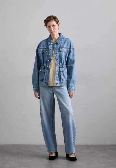 Gai zilā denim jaka ar izsvērta vidukļa līniju, četriem kabatām un pogu aizdari, valkāta virs krēmkrāsas acu adījuma džempera un gaišiem džinsiem.