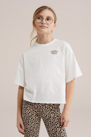 Jeune personne portant des lunettes et un T-shirt blanc oversize avec le texte "GOLDEN HOUR" ainsi que des leggings à imprimé léopard, debout devant un fond uni.