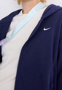 Marineblauer Hoodie mit Reißverschluss und weißem Nike-Logo, getragen über einem hellen Shirt. Ein hellblauer Gurt mit einem Clip hängt aus dem Halsbereich.