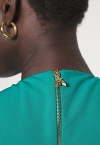 Tyrkysové šaty s pozlaceným zipem na zadní straně a charakteristickým zipem ve tvaru listu. Hladká textilie a přiléhavý střih.