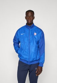 Modrá bunda Nike s textúrovaným vzorom z opakujúcich sa písmen, predným zapínaním na zips a malým logom na ľavej strane hrudníka.