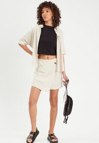 Shorts en lin beige avec un design enroulé et un détail de bouton, associés à un haut noir ajusté court et des sandales noires, tenant un petit sac noir.
