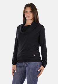 Maglione nero a collo alto realizzato in tessuto morbido, con maniche lunghe e dettaglio del logo in basso, abbinato a jeans blu.