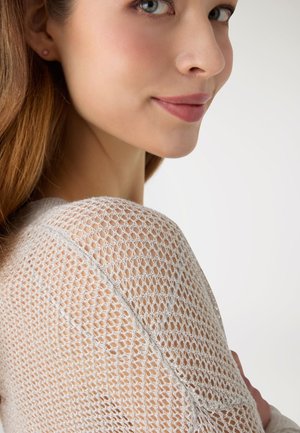 Donna con capelli castano chiaro indossa un maglione bianco a maglia aperta, mostrando un primo piano del suo volto e della spalla su uno sfondo uniforme.