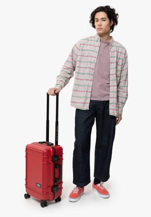 Red hard-shell suitcase with retractable handle and wheels, textured surface, and contrasting black hardware; person holds it while wearing casual attire.