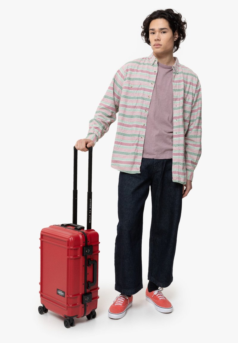 Red hard-shell suitcase with retractable handle and wheels, textured surface, and contrasting black hardware; person holds it while wearing casual attire.