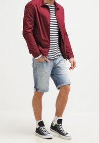 Rode lichte jas, marineblauw en wit gestreept shirt, knielange lichtblauwe denimshorts, zwarte hoge sneakers met witte accenten.