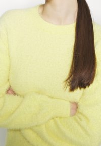 Żółty, puszysty sweter z dzianiny o okrągłym dekolcie i długich rękawach. Miękka tekstura, dopasowane ściągacze, ramiona skrzyżowane z przodu.