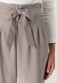 Pantalons taille haute à carreaux en beige, marron et bleu marine avec un détail nœud à la taille et des plis, fabriqués à partir d'un tissu texturé.
