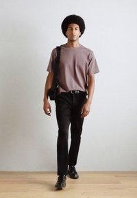 Un tricou gri, pantaloni mulați negri și pantofi negri. Modelul poartă o geantă messenger neagră, cu un podea din lemn și un fundal simplu.
