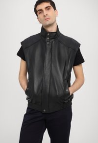 Μαύρο δερμάτινο γιλέκο χωρίς μανίκια με ψηλό γιακά, κουμπιά με κούμπωμα και πλαϊνές τσέπες. Λιτή υφή και εφαρμοστή σχεδίαση με σ�υγκεντρωμένη ραφή στο κάτω μέρος.