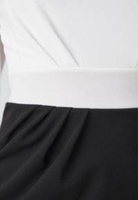 Čierno-biele šaty s priliehavým telom, ktoré majú plisovanie v čiernom materiáli a hladký biely pás. Celkový mäkký materiál.