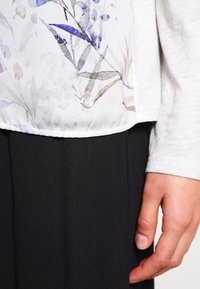 Tessuto bianco con motivo floreale, accentuato da tonalità di blu e beige, caratterizzato da una texture liscia e una chiusura con bottone, su uno sfondo di materiale nero.