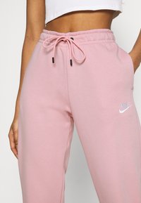 Pink sweatpants with a drawstring waistband, featuring black accent hardware and a white Nike logo. Made from soft, textured fabric.