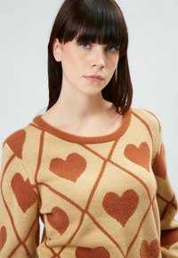 Beigefarbener Strickpullover mit braunem Herzmuster. Er hat einen runden Ausschnitt und eine weiche Textur, mit einem Gitterdesign, das die Herzen betont.