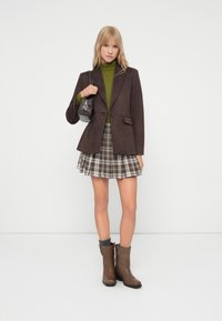 Bruine blazer, groene coltrui, geruite plissérok en bruine enkellaarsjes. Accessoires omvatten een kleine grijze handtas. Efficiënte texturen en een op maat gemaakte pasvorm.