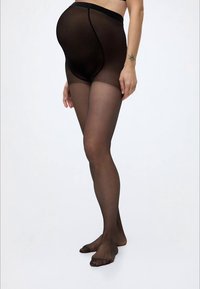 Personne enceinte portant des collants de maternité noirs transparents, debout pieds nus devant un fond clair uni.