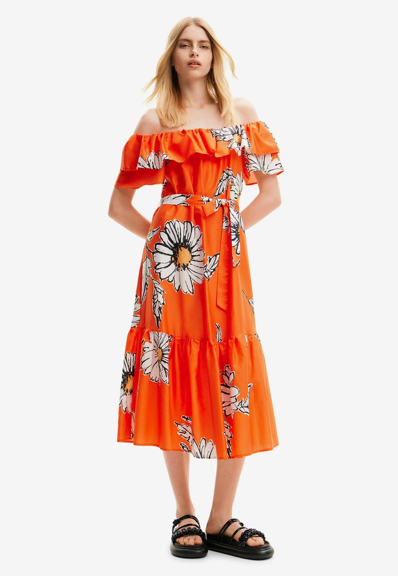 Schouderloos oranje jurk met bloemenprint, ruches, stoffen riem en gelaagd ontwerp. Gecombineerd met zwarte sandalen.