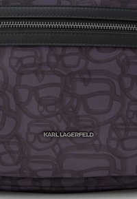 Ciemnofioletowa torba z tkaniny z abstrakcyjnym wzorem twarzy, czarny zamek błyskawiczny oraz metalowe logo marki "KARL LAGERFELD" umieszczone centralnie pod zamkiem.