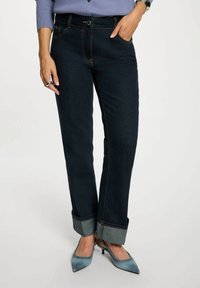 Jeans in denim blu scuro con vestibilità affusolata, caratterizzati da un risvolto, cuciture a contrasto e tasche anteriori. Indossati con scarpe con tacco blu.