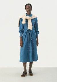 Denim-mekko, jossa on vyötärönauha, pitkät hihat ja nappilista etupuolella. Päällä vaaleanruskea neulottu villapaita, joka on laskettu hartioille.