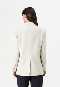 Beige tilpasset blazer med notched krage, enkelt ventil bak og knapper på mansjettene. Glatt tekstur og strukturert passform.