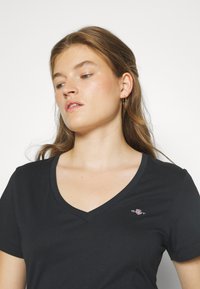 Mlada žena valovite svijetlosmeđe kose koja nosi zlatne naušnice u obliku obruča i crnu T-majicu s V-izrezom s malim logotipom na lijevoj strani prsa, gleda u stranu.