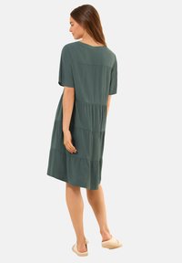 Vihreä kerroksellinen mekko, joka on valmistettu kevyestä kankaasta. Lyhyet hihat, pyöreä pääntie ja rento leikkaus. Vaaleat liukuvaatteet.