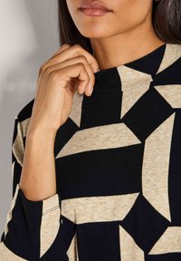 Eine Frau, die den Kragen eines langärmeligen Shirts mit schwarz-beigem geometrischem Muster anpasst, wobei ein Teil ihres Gesichts und ihrer Hand sichtbar ist.