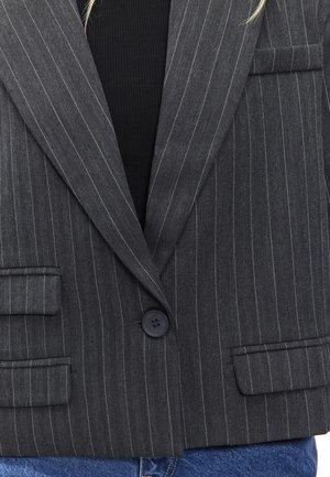 Blazer gris à rayures avec une texture en chevrons, fermeture à un bouton, et deux poches avant. Porte sur un haut noir et un denim.