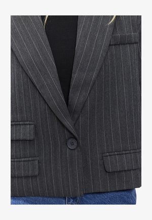 Blazer gris à rayures avec une texture en chevrons, fermeture à un bouton, et deux poches avant. Porte sur un haut noir et un denim.