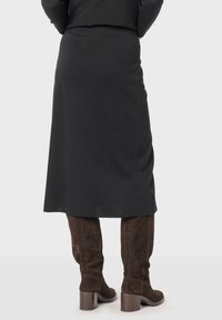 Jupe midi noire en tissu doux, avec une texture lisse et un léger drapé sur le côté, associée à de hautes bottes en daim marron.