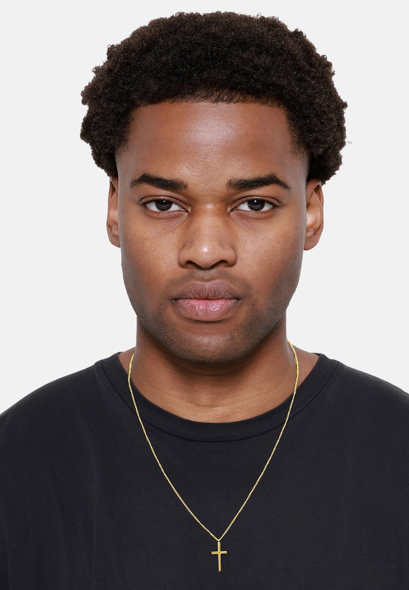 Zwarte T-shirt, krullend zwart haar en een gouden ketting met een klein kruisje als hanger. Effen achtergrond zonder extra elementen.