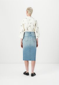 Persoană cu păr scurt blond, purtând o bluză albă cu imprimeu floral, fustă de denim albastru deschis până la genunchi și pantofi negri, stând cu spatele la privitor.