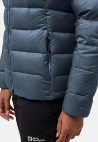 Blaue gepolsterte Jacke mit glatter Textur, horizontalen Nähten, einer Reißverschlusstasche und einer engen Passform. Schwarze Hosen mit einem Logo.