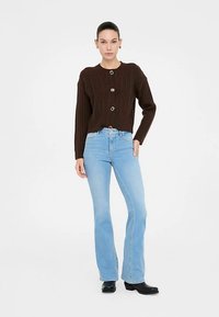 Barna, kötött kardigán bordázott ujjakkal, díszgombokkal. Világoskék, szárában bővülő farmerral és fekete bokacsizmával viselve.