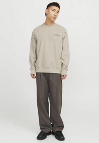 Beige pulóver bordázott mandzsettákkal és szegéllyel, elöl fekete szöveggel. Laid-back sötétszürke nadrággal és fekete cipővel párosítva.