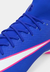 Μπλε ποδοσφαιρικό παπούτσι με υφή στην επάνω πλευρά, λευκές και ροζ λεπτομέρειες και μπλε κορδόνια. Κομψός σχεδιασμός με αεροδυναμικό σχήμα.