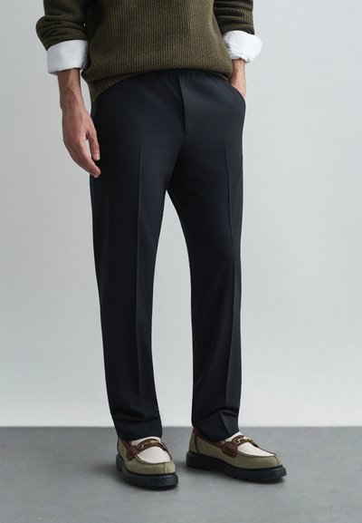 Black tailored pants with a smooth texture, paired with multicolored loafers featuring a brown accent and white detailing.