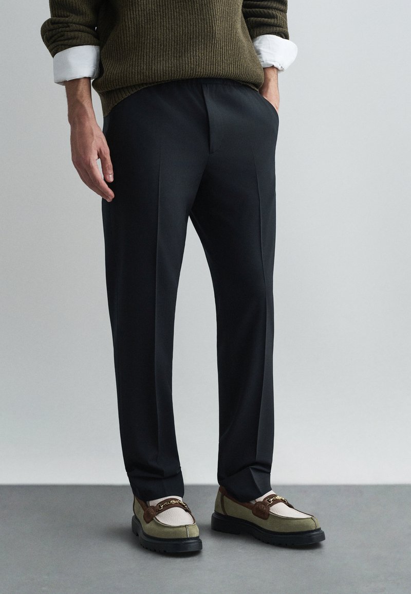 Juodos pritaikytos kelnės su lygia tekstūra, derinamos su daugiaspalviais loaferiais, turinčiais rudą akcentą ir baltus akcentus.