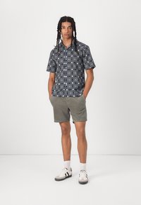 Chemise à manches courtes à motifs noir et blanc associée à un short vert olive et des baskets blanches avec des accents noirs. Design simple, matériau léger.