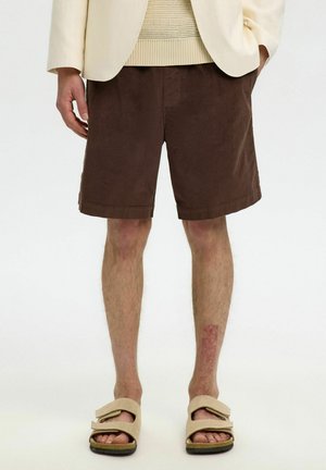 Man draagt bruine corduroy korte broek, beige sandalen met dubbele band, crèmekleurige gebreide trui en lichtbeige blazer, staat met handen in de zakken.