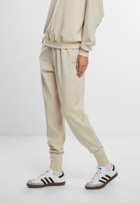 Beige hettegenser og matchende joggebukser i myk stoff, med ribbestrikkede mansjetter og sidelommer. Kombinert med hvite og svarte joggesko.